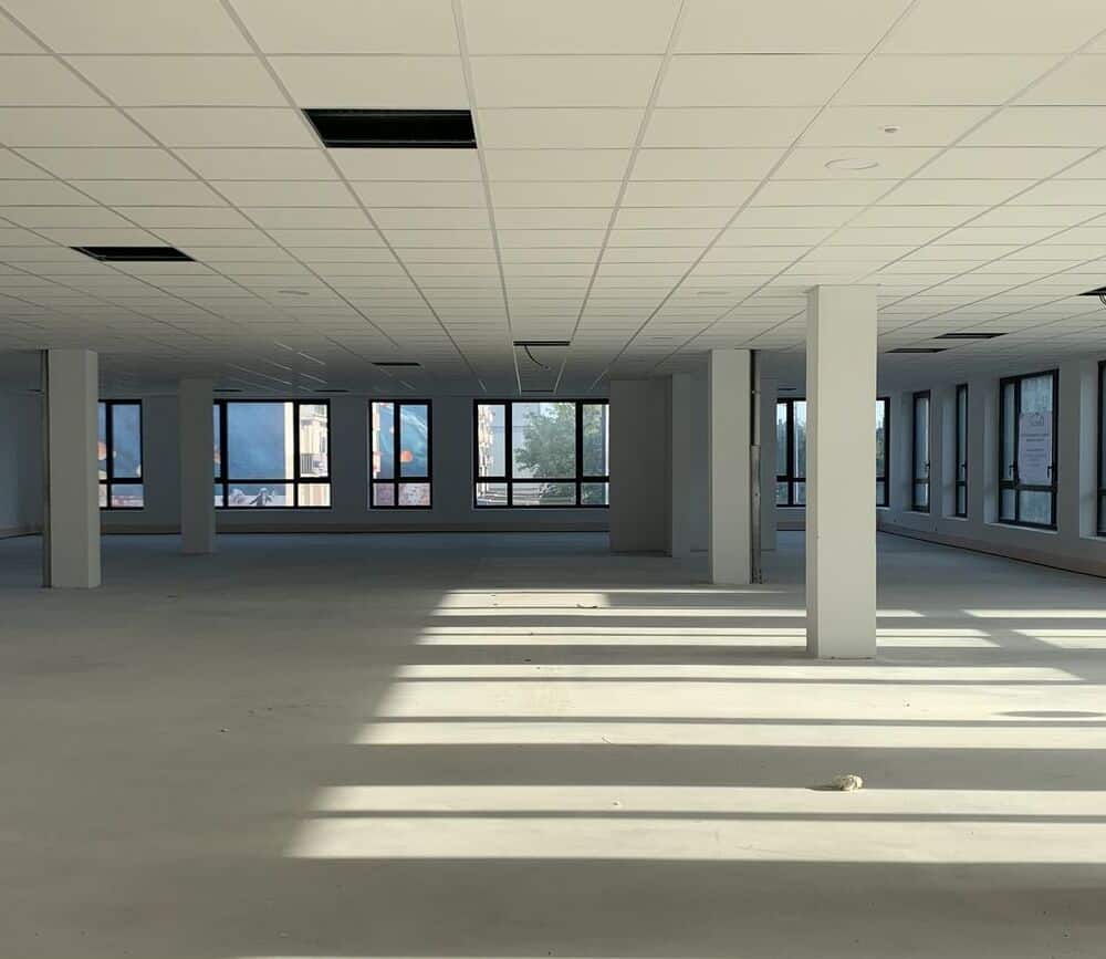 Open space bureau vide : locaux commerciaux en attente d'aménagement. Vaste open space de bureaux en construction, sol nu et faux plafond blanc. Les rayons du soleil entrant par les fenêtres projettent de longues ombres dramatiques.